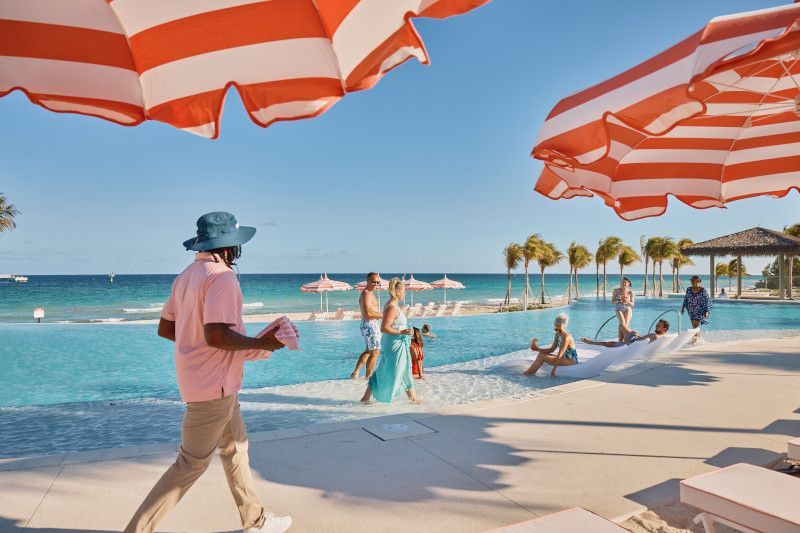  Guests enjoy a sunny day at Celebration Key's oceanfront infinity pool, featuring white and orange striped umbrellas, palm trees, and a floating hammock lounge.