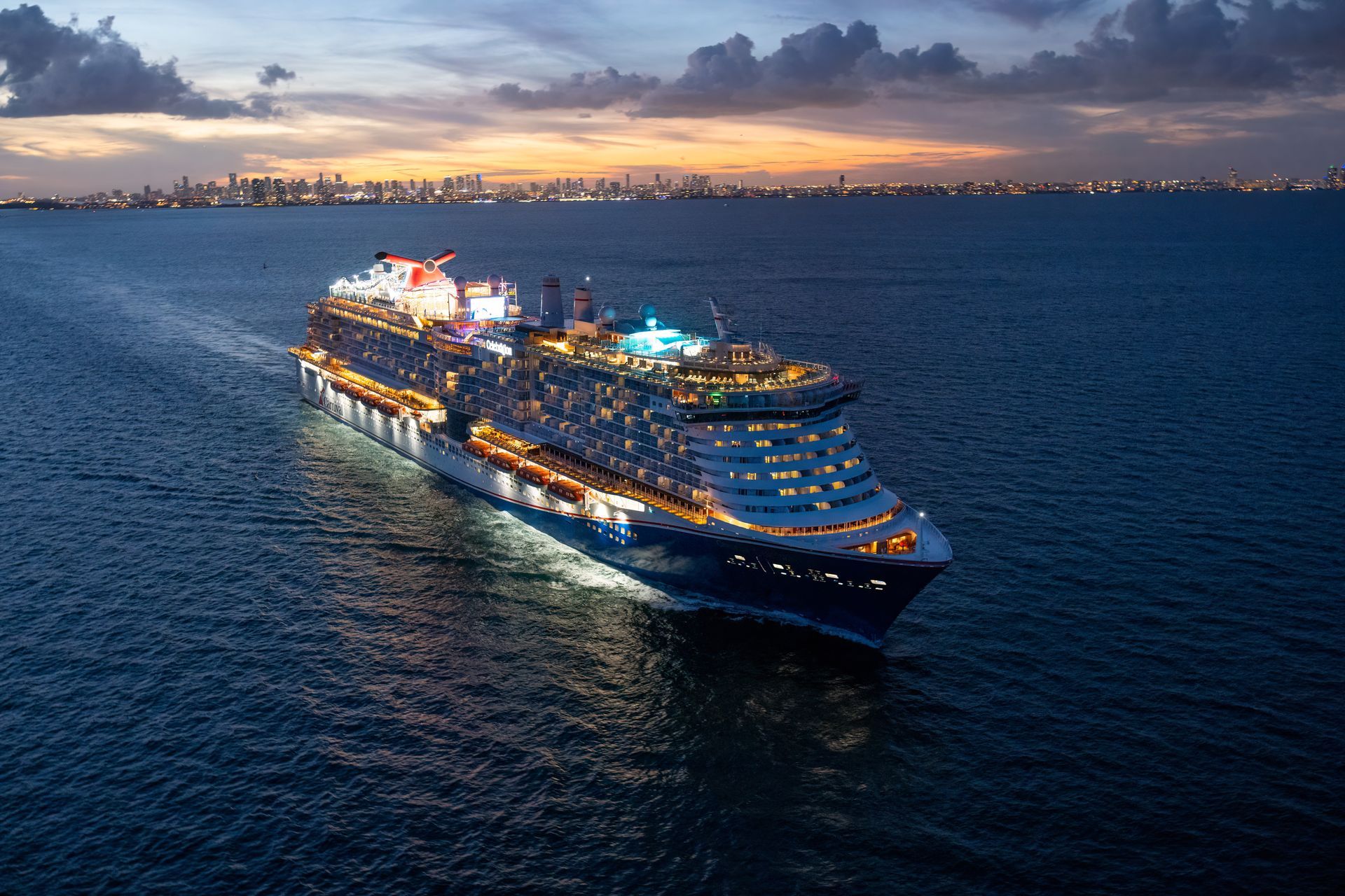 Cruise ship sailing on dark blue water at dusk with city skyline in the distance.
