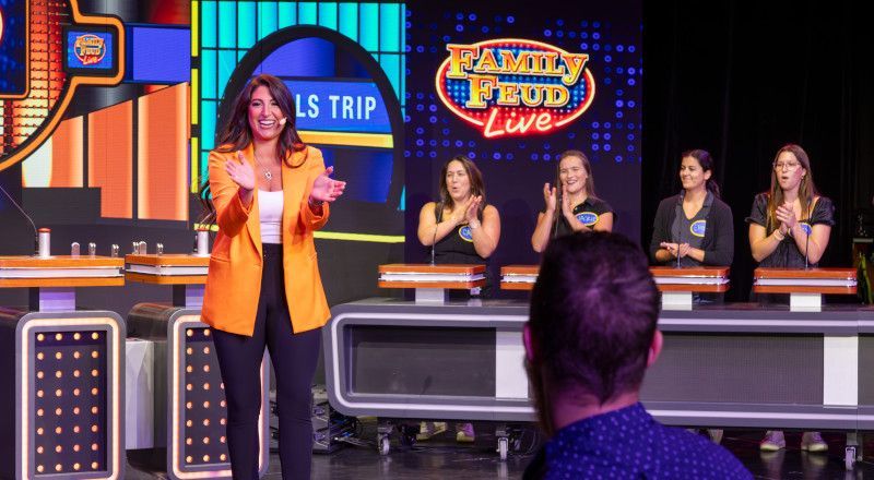 A woman in orange blazer clapping on a Family Feud stage with a group of women applauding.