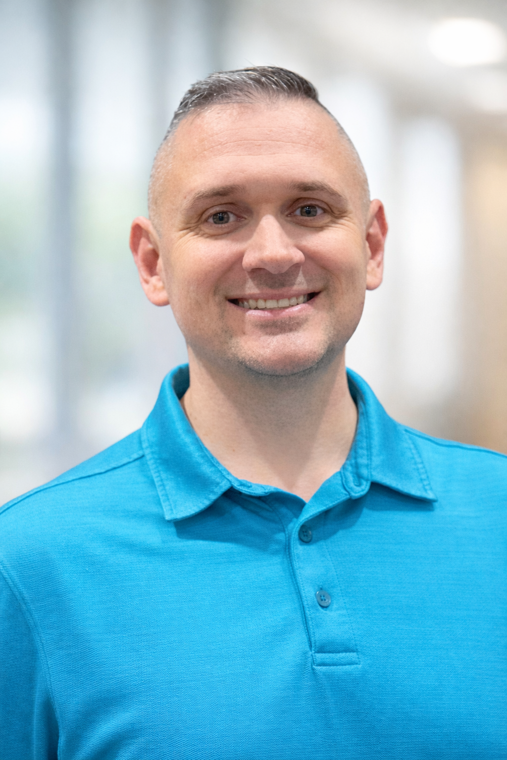 Smiling man in a bright blue polo shirt standing indoors against a blurred office background