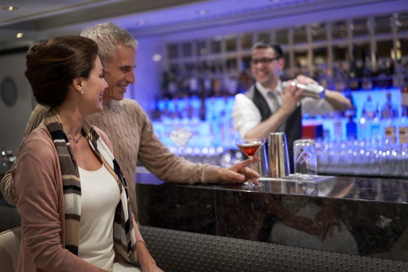 Couple at bar watching bartender mixing a drink; blue backlighting, elegant setting.