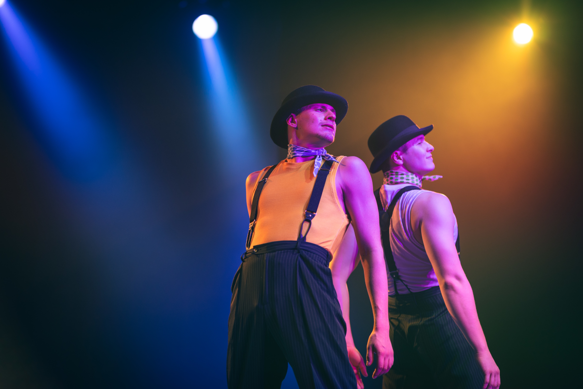 Two dancers in hats and suspenders under stage lights. One in yellow, one in white.