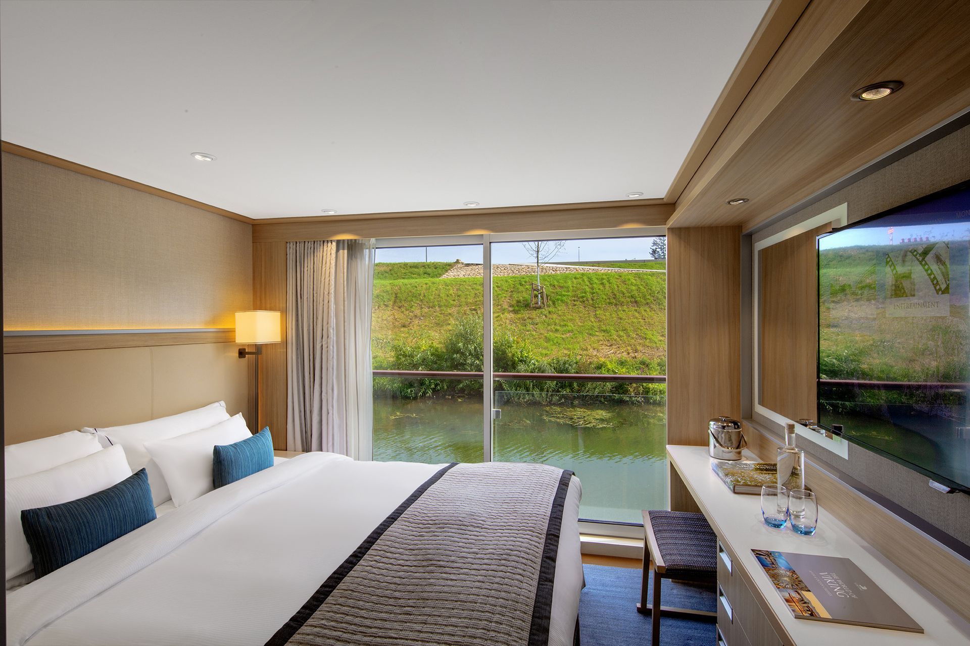 Bedroom on a river cruise ship with a large window overlooking green foliage and water. The room has a bed, desk, and TV.