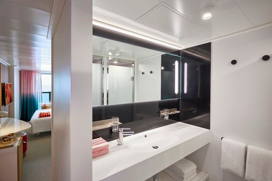 Modern bathroom interior with a large mirror, white sink, and towel rack. A bedroom is visible in the background.
