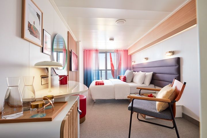 Interior view of a Virgin Voyages Sea Terrace cabin with a queen bed, stylish mid-century décor, desk area, and floor-to-ceiling doors leading to a balcony with a red hammock.