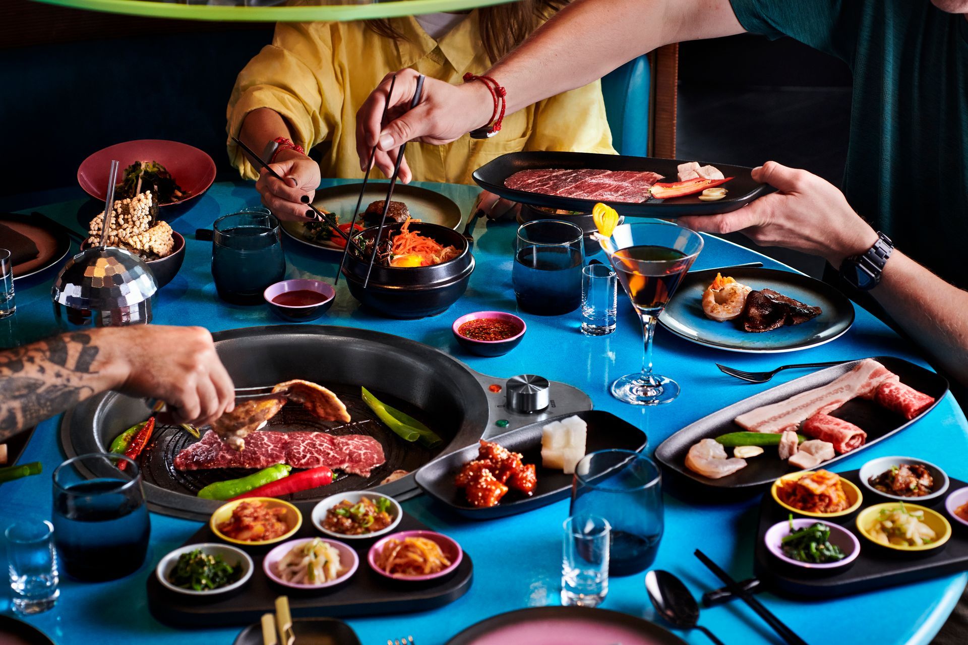 People eating Korean barbecue at a blue table, grilling meat with side dishes and cocktails at Gunbae - Virgin Voyages' dedicated Korean BBQ restaurant
