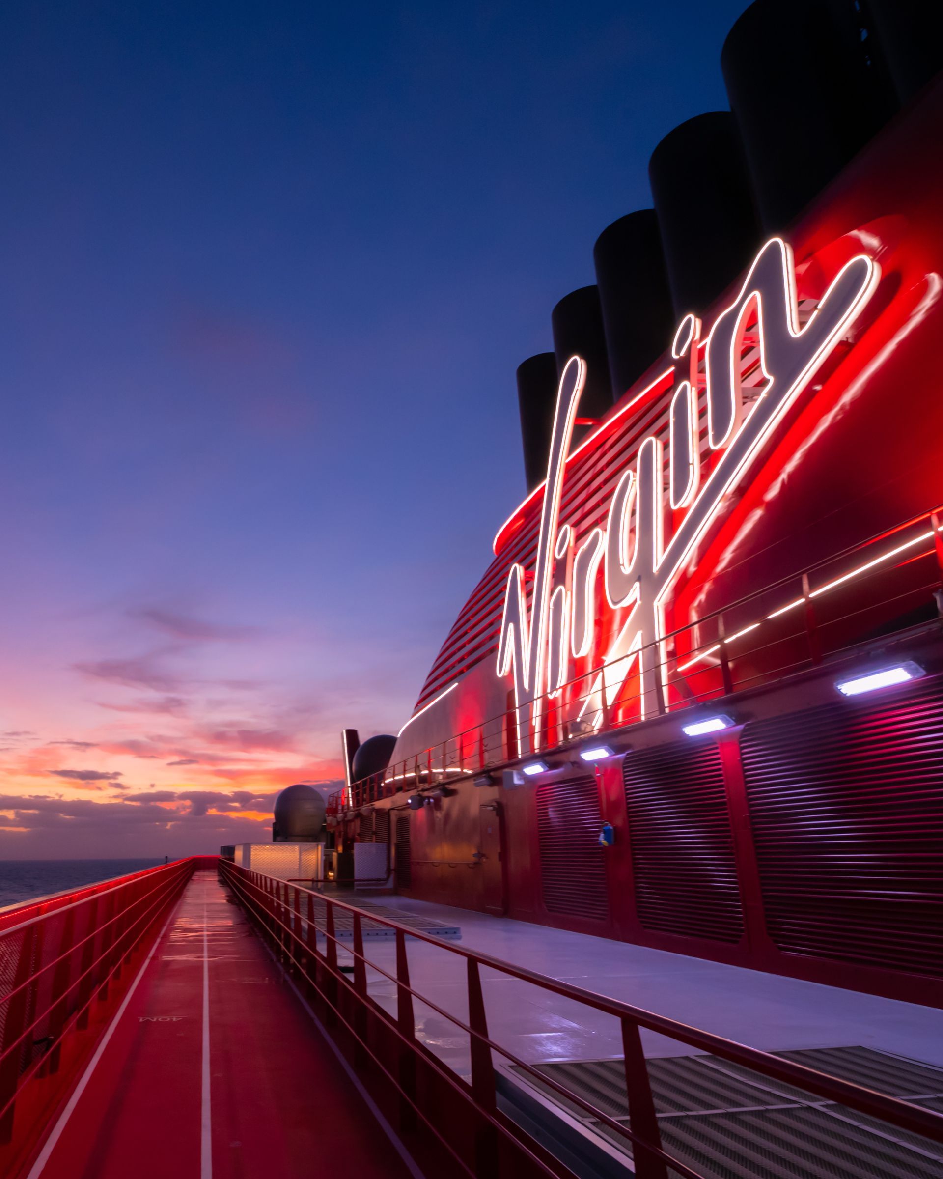 Close-up of the illuminated Virgin Voyages logo glowing in red on the ship’s deck at sunset, with the ocean and colorful evening sky in the background.
