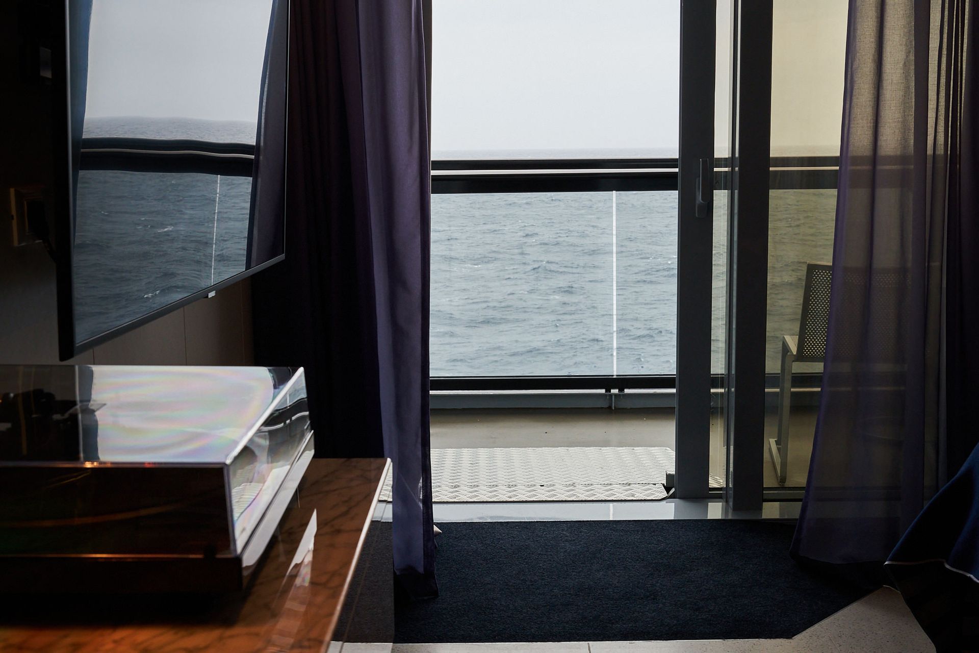 Interior view of room with balcony and ocean view. Dark curtains frame a glass door and railing.
