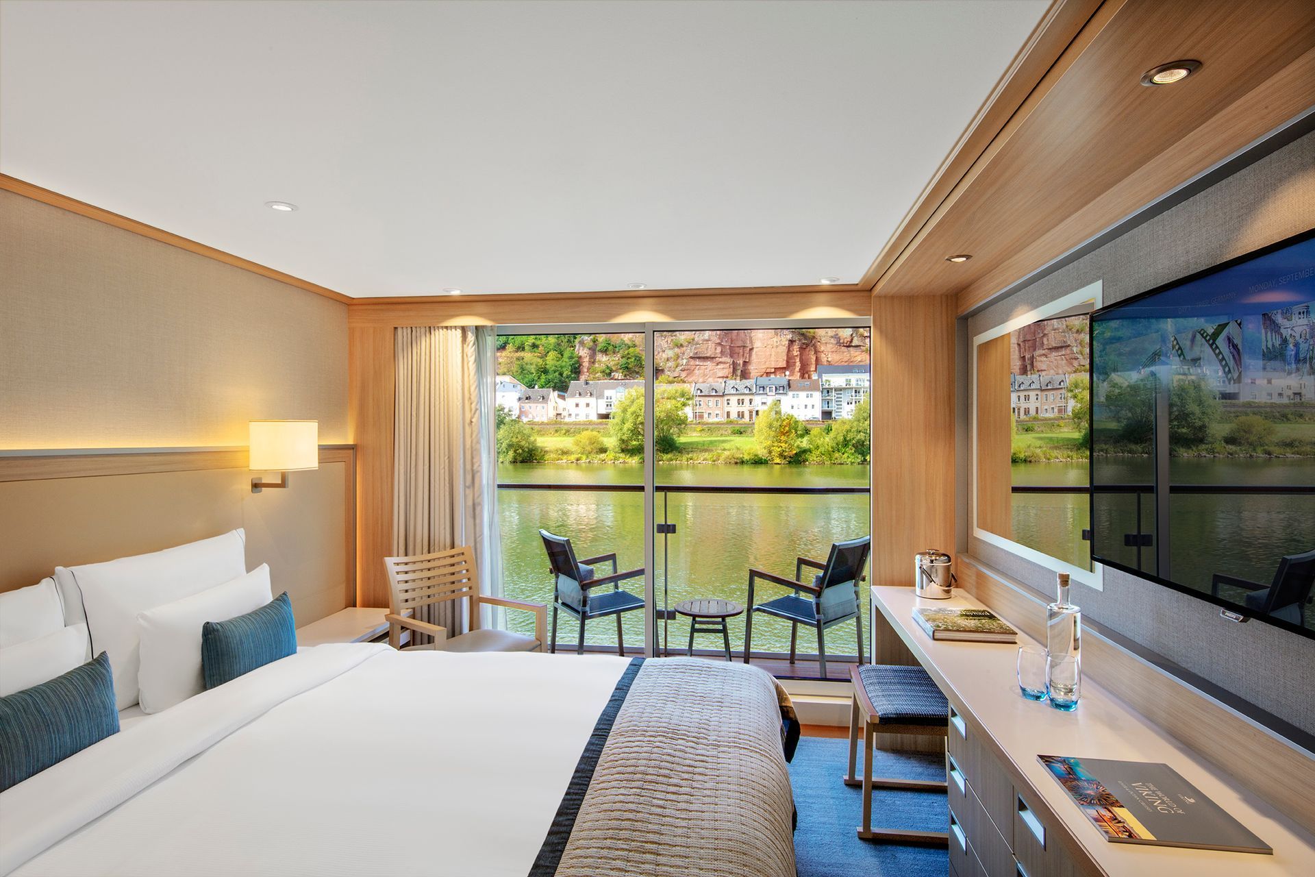 Bedroom on a river cruise ship, with a bed, balcony, and view of a river and town.