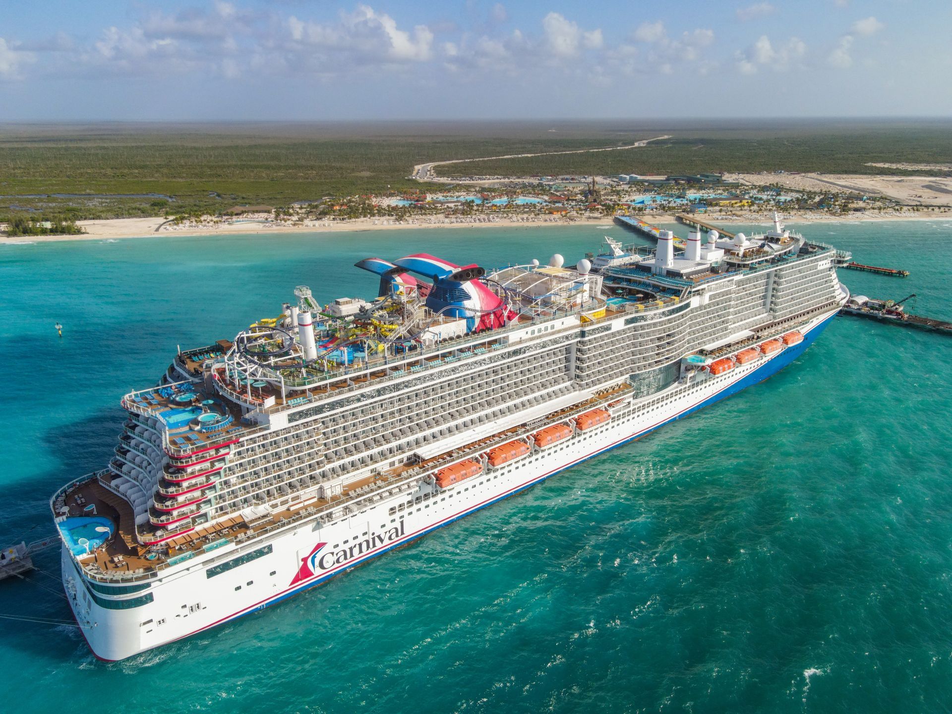 Large Carnival cruise ship docked at a tropical beach. The ship is white, red, and blue.