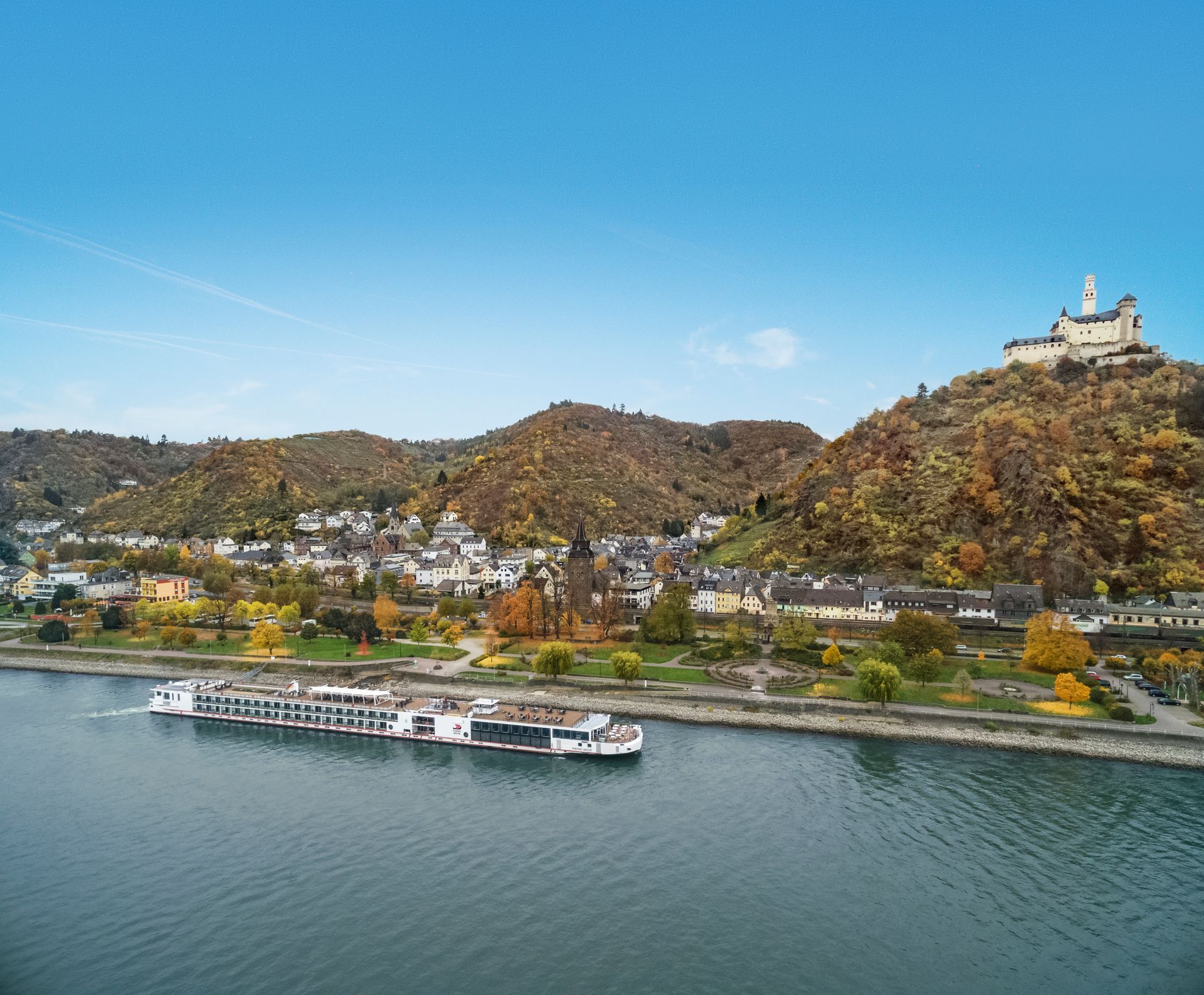 A white river cruise ship sailing past a grand medieval castle on a sunny day with lush greenery and clear reflections on the water.