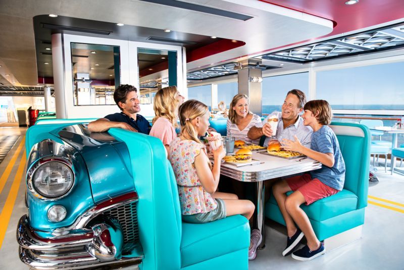 Family dining at a retro diner booth with a car front, blue seats, and ocean view.