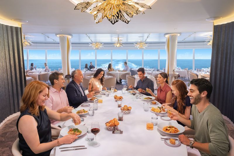 A group dines at a long table in a ship's dining room with ocean views; smiles, food, and joy are visible.