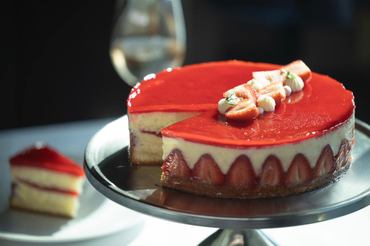Strawberry cake on a silver platter with a slice on a plate; glazed top, berries around the base.