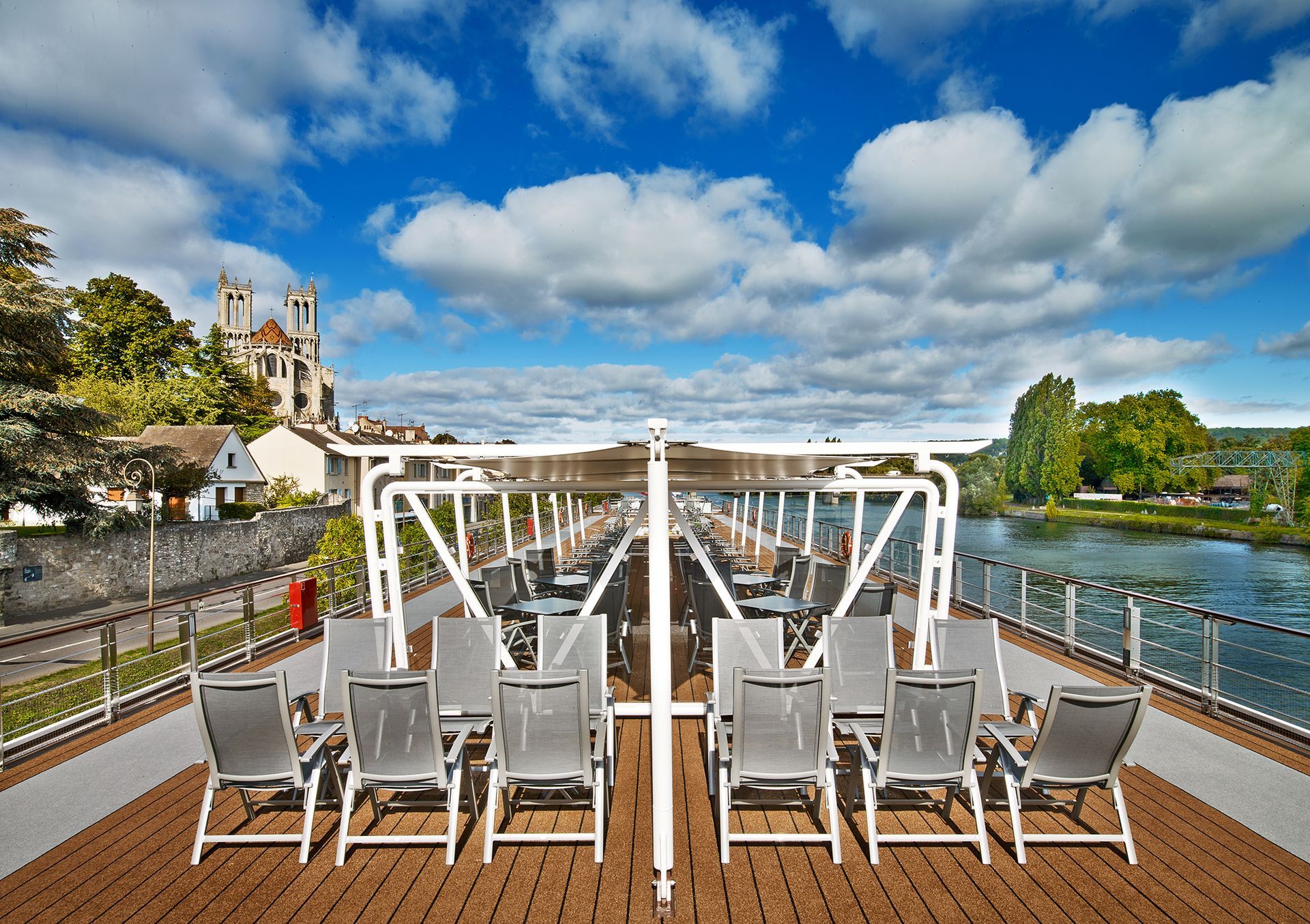 Deck of a river cruise ship with lounge chairs, overlooking a river and a building with a tower under a blue sky.