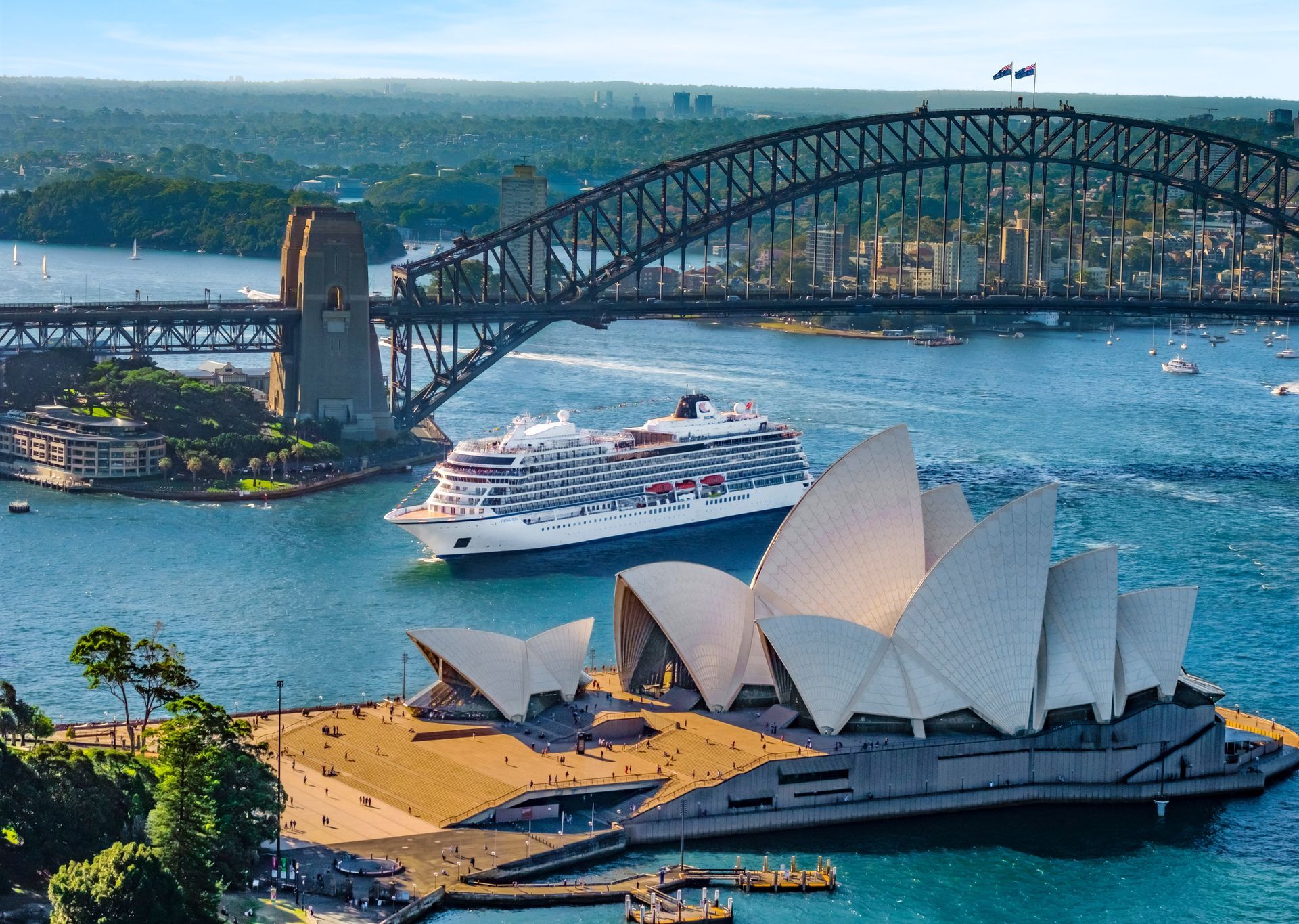 Aerial view of a Viking ocean ship in Sydney Harbour, sailing between the Sydney Opera House and the Harbour Bridge on a sunny day.