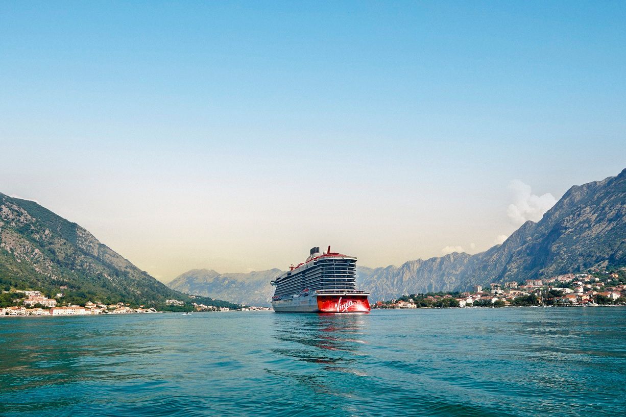 Virgin Voyages cruise ship sailing through calm blue waters surrounded by coastal villages and mountain ranges of Montenegro, showcasing the line’s scenic Mediterranean-style itinerary and modern ship design.