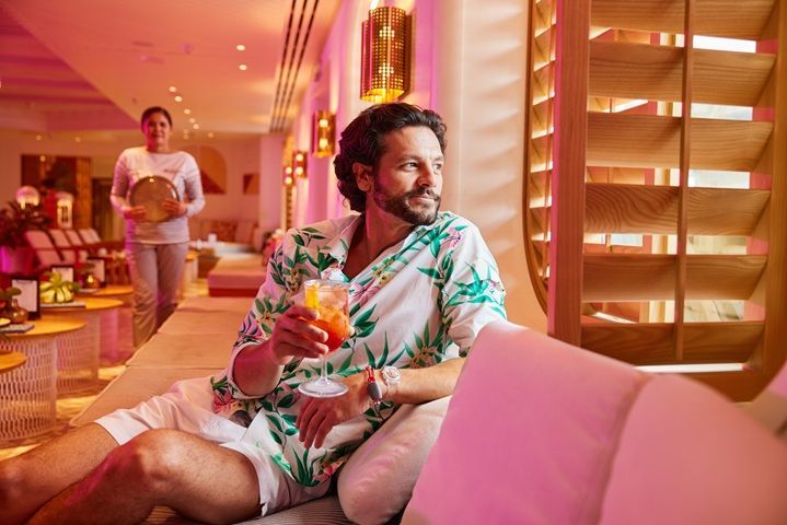 A guest relaxes indoors on a Virgin Voyages ship, enjoying a colorful cocktail in a lounge with warm lighting and modern décor as a crew member approaches with a tray.