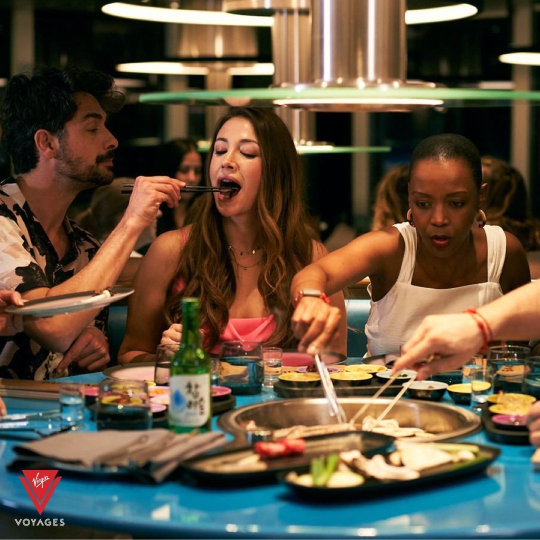People at a restaurant, eating. One person feeds another. Bright, colorful, blue table at Gunbae - Virgin Voyages' dedicated Korean BBQ restaurant.