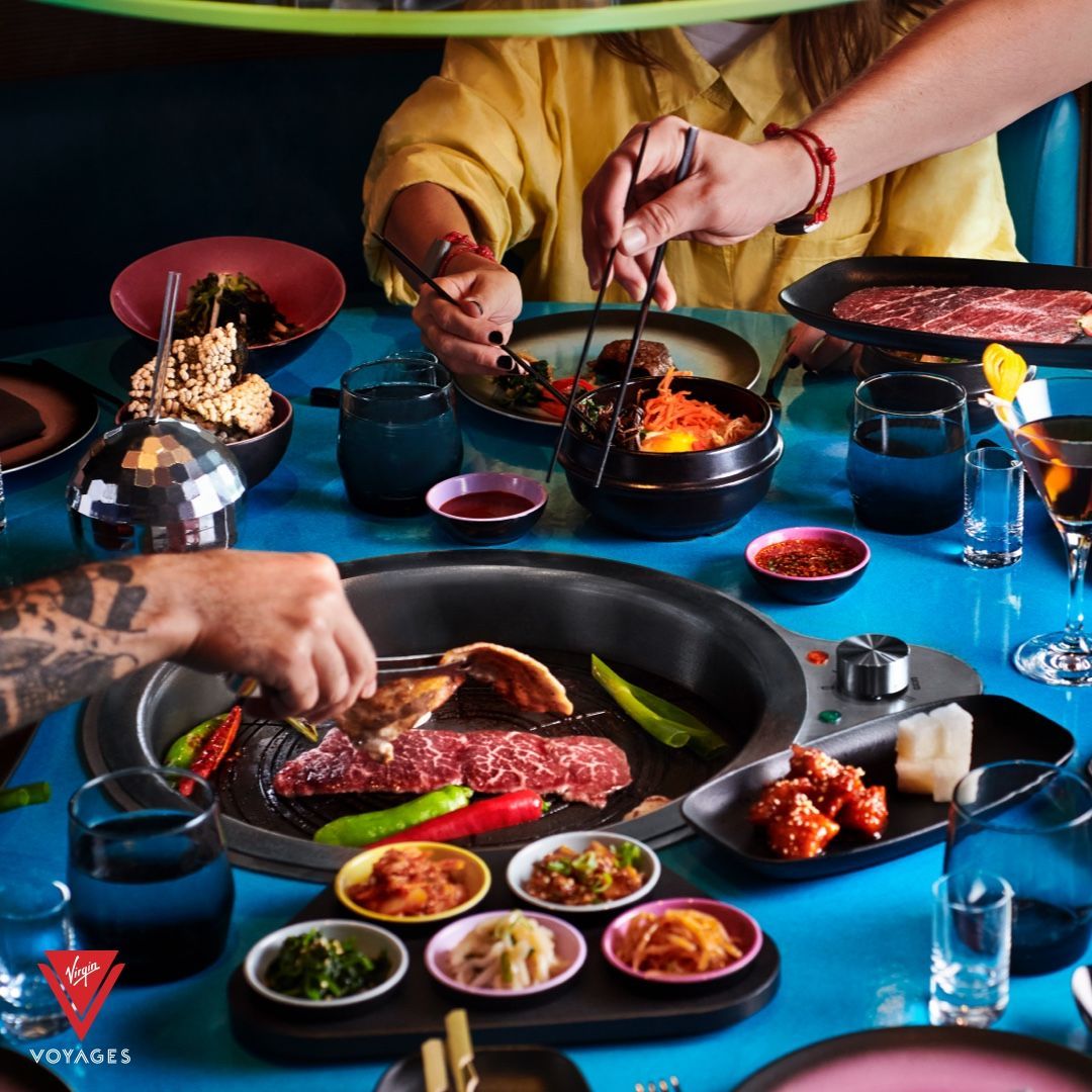 People at a table grilling Korean food. Dishes of food and drinks on a bright blue table at Gunbae - Virgin Voyages' dedicated Korean BBQ restaurant.
