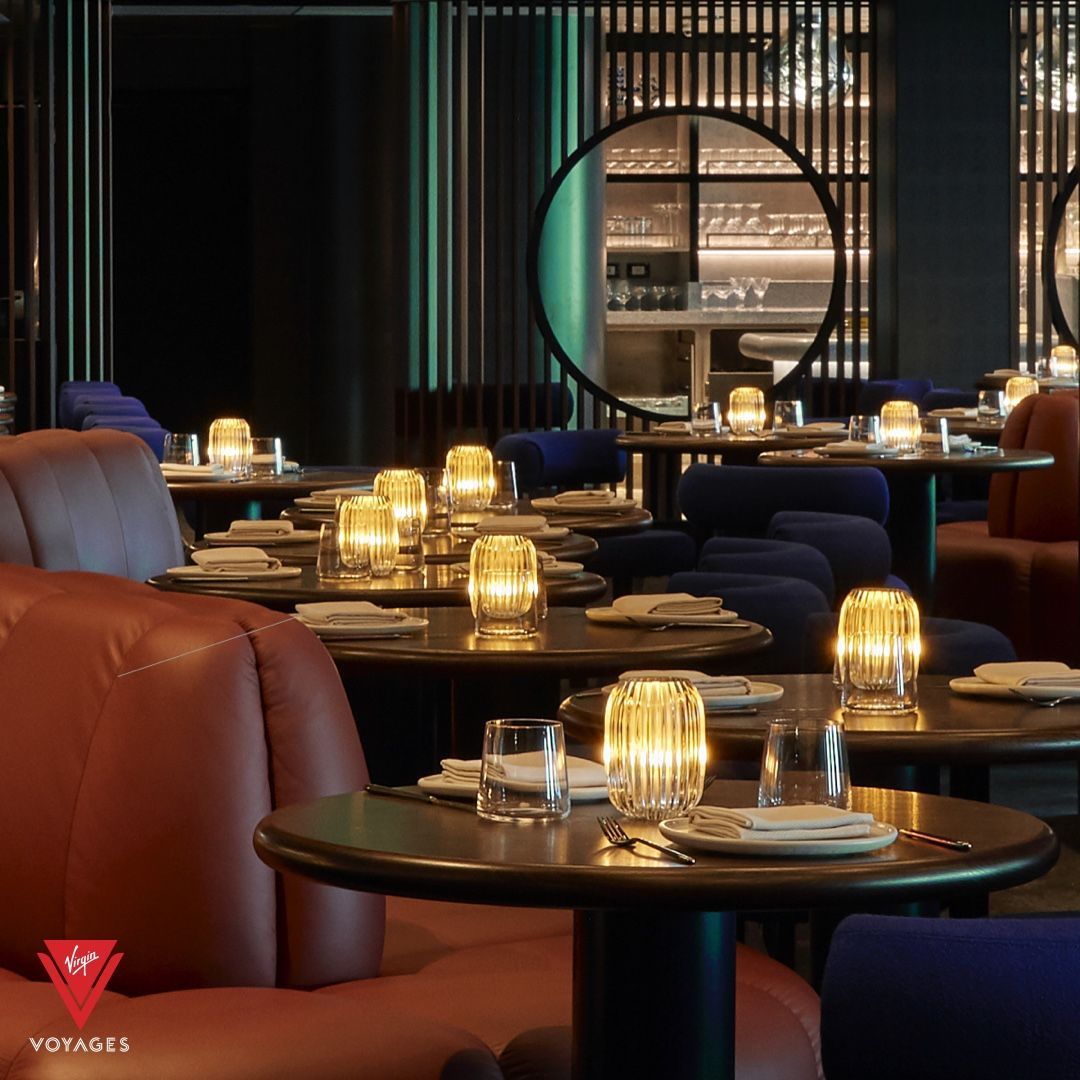 Interior of a restaurant with round tables, soft lighting, and red and blue seating at Pink Agave - Virgin Voyages modern Mexican restaurant.