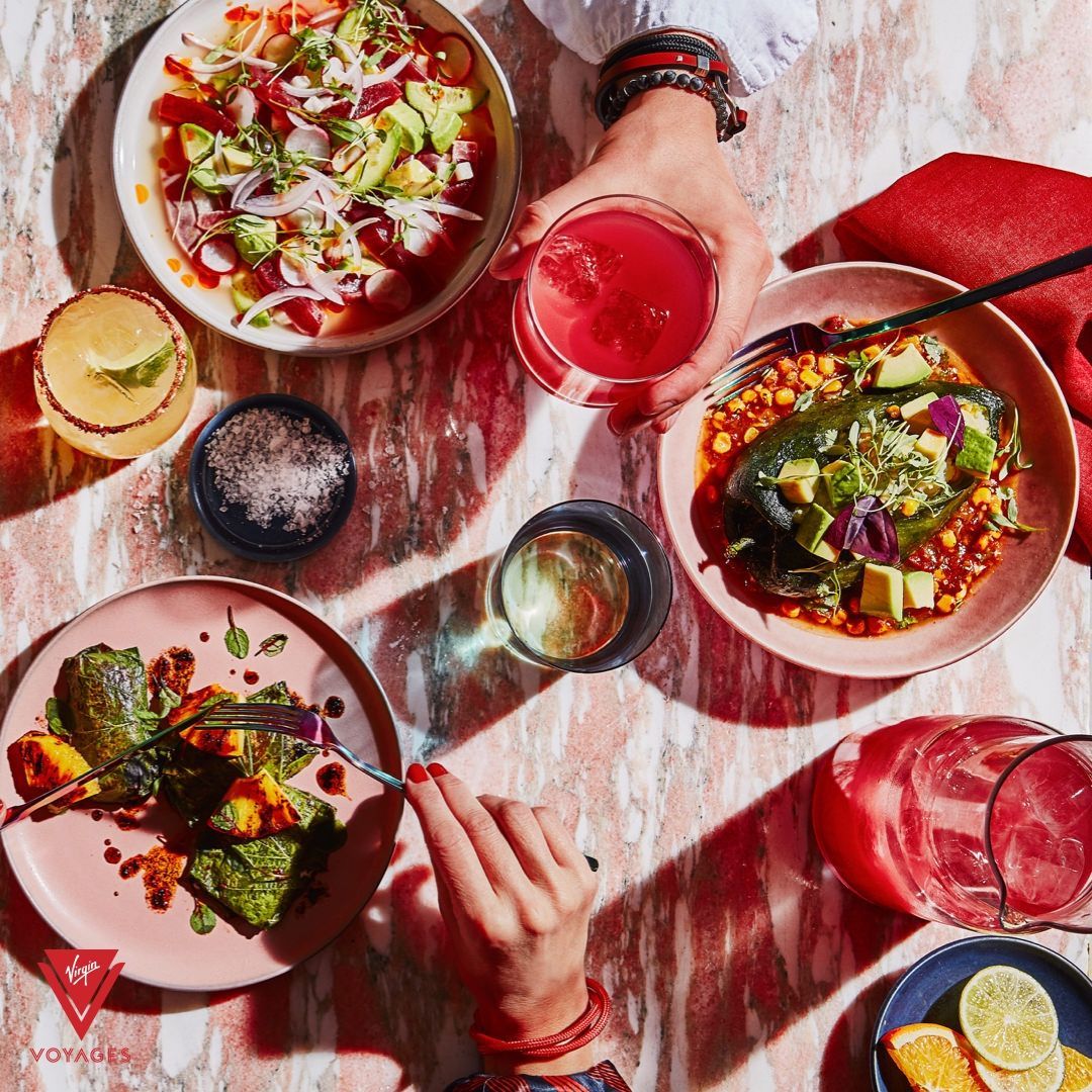Overhead shot of a table with Mexican food and drinks, hands reaching for a glass and a plate at Pink Agave - Virgin Voyages modern Mexican restaurant.