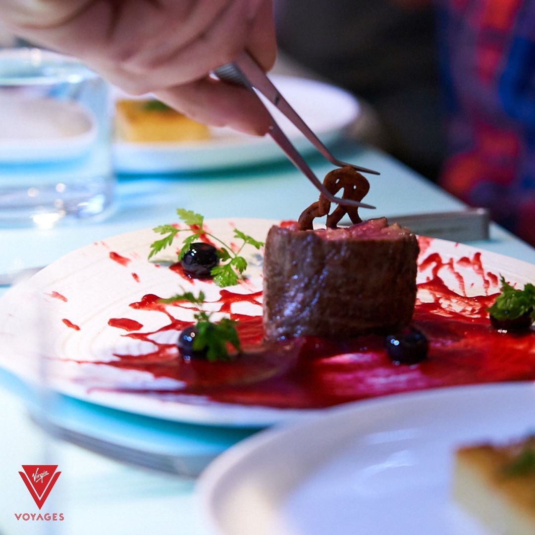 Chef using tongs to garnish a steak with a pretzel-like element. Red sauce and blueberries on the plate at The Test Kitchen - Virgin Voyages Experimental, chef-led tasting lab serving inventive, multi-course dishes.