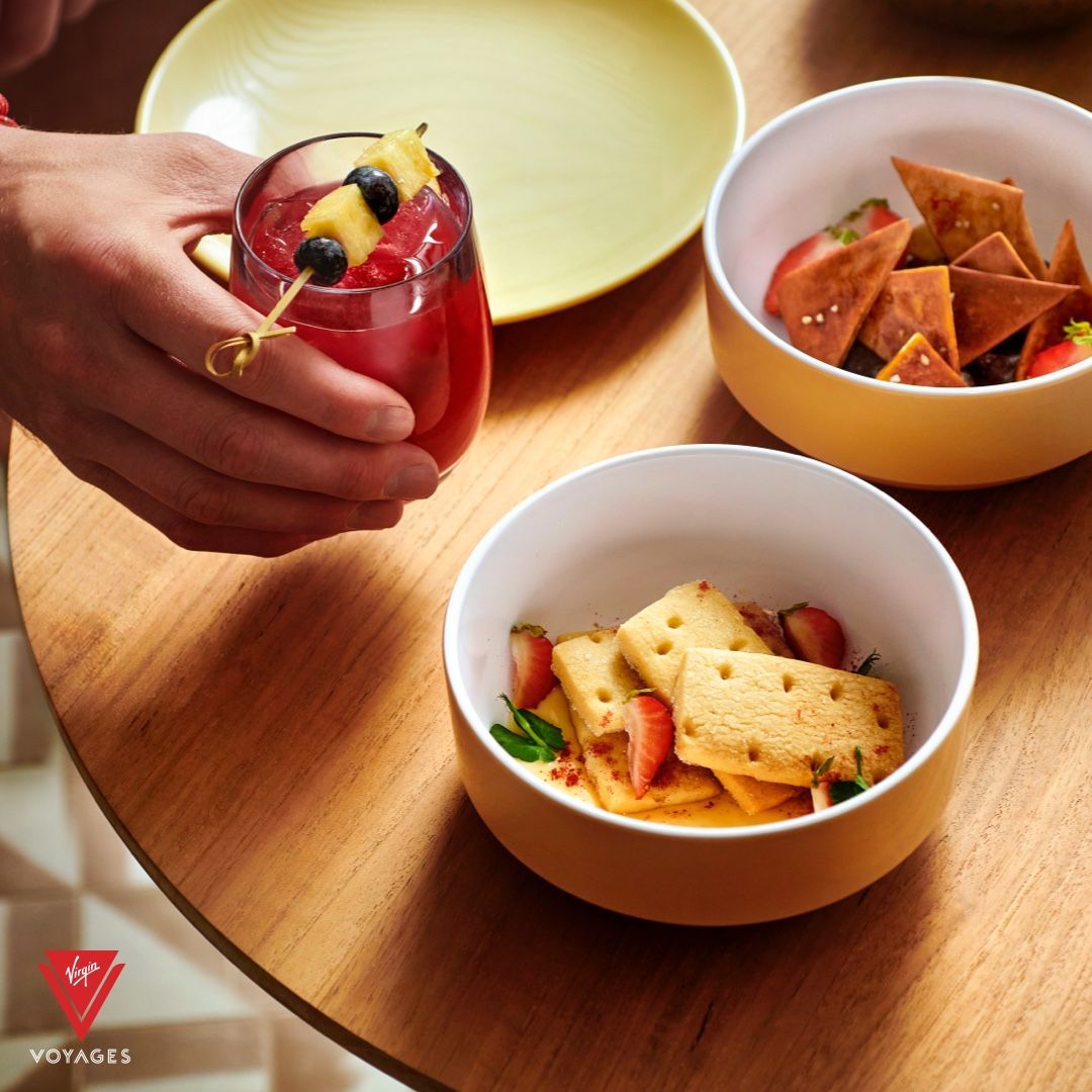 Person holding a red drink with fruit on a skewer, with bowls of food on a table at The Dock - Virgin Voyages' dedicated Mediterranean-inspired open-air and indoor lounges.