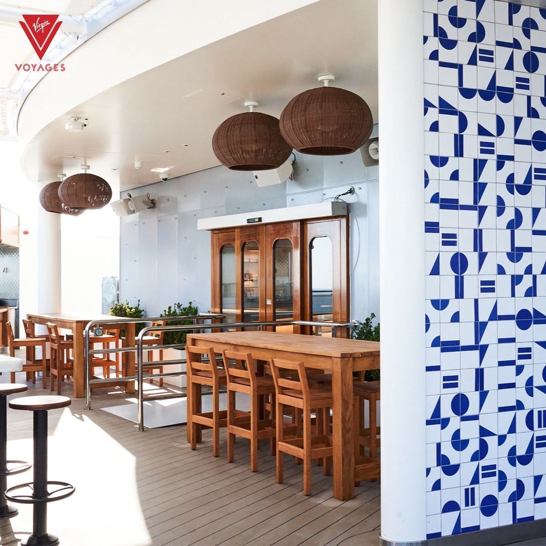 Outdoor bar area with wooden tables and chairs, white walls, and blue and white tile design at The Dock - Virgin Voyages' dedicated Mediterranean-inspired open-air and indoor lounges.