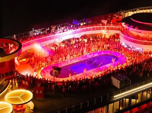 Guests celebrating at a Virgin Voyages pool deck party illuminated by red and purple lights, reflecting the cruise line’s vibrant adults-only nightlife and entertainment experience.
