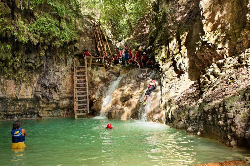 People in turquoise water at a waterfall with rock walls and wooden stairs.