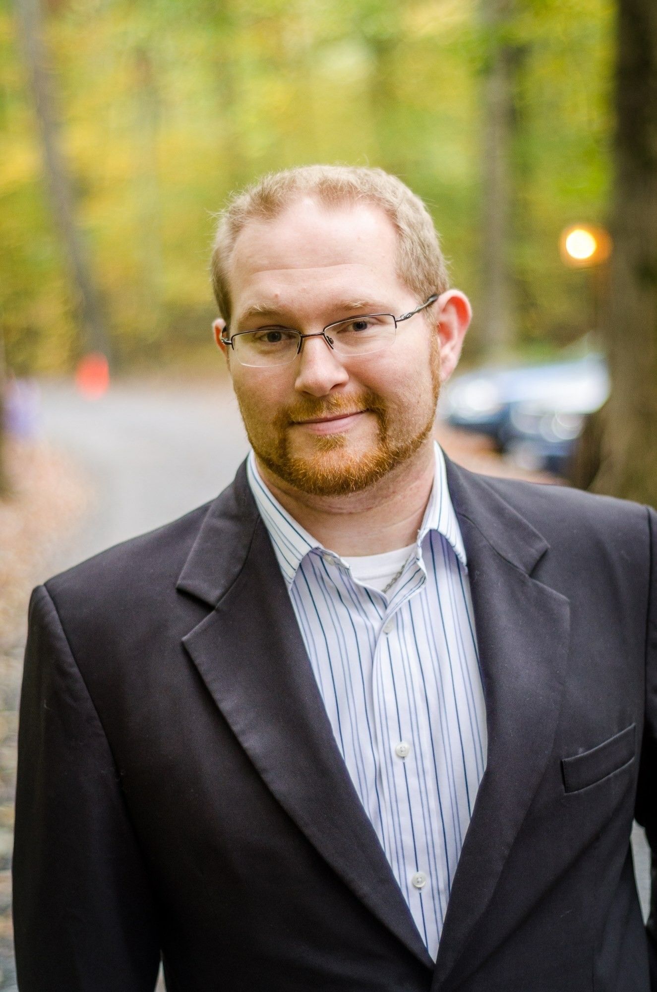 Man in a dark blazer and striped shirt standing outdoors, smiling softly against a blurred autumn background