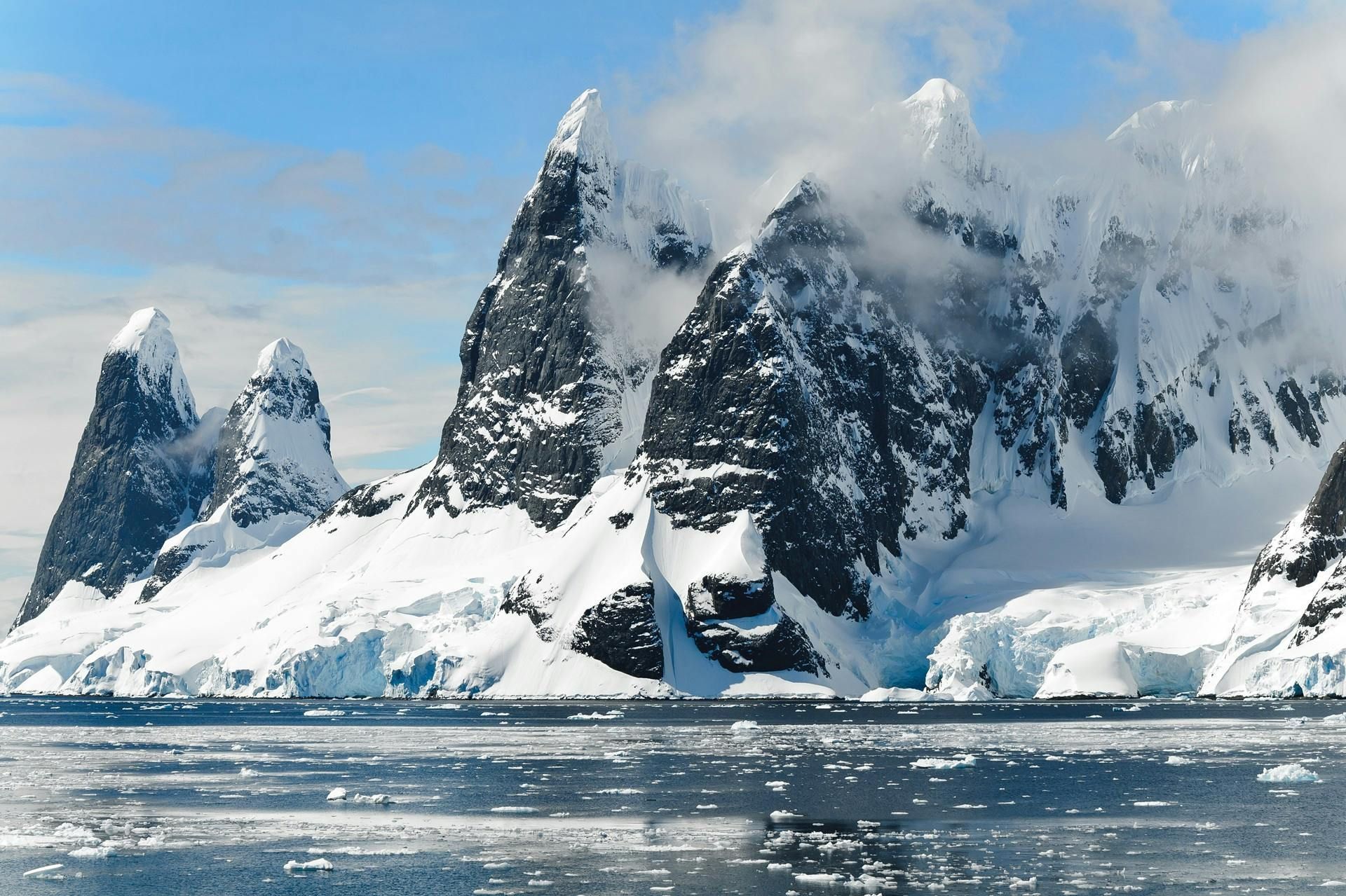 Snow-covered mountains in Antarctica with clouds, a blue sky, and icy sea.