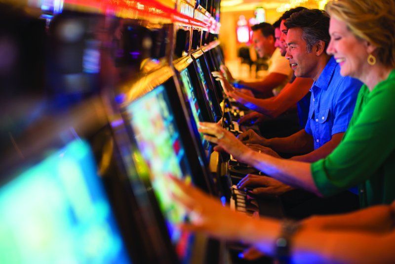 People playing slot machines in a casino. Focus on hands on screens; bright lights and smiling faces.