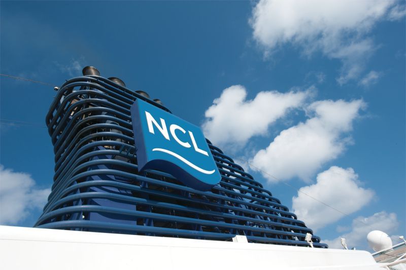 Blue NCL cruise ship smokestack against a bright blue sky with white clouds.