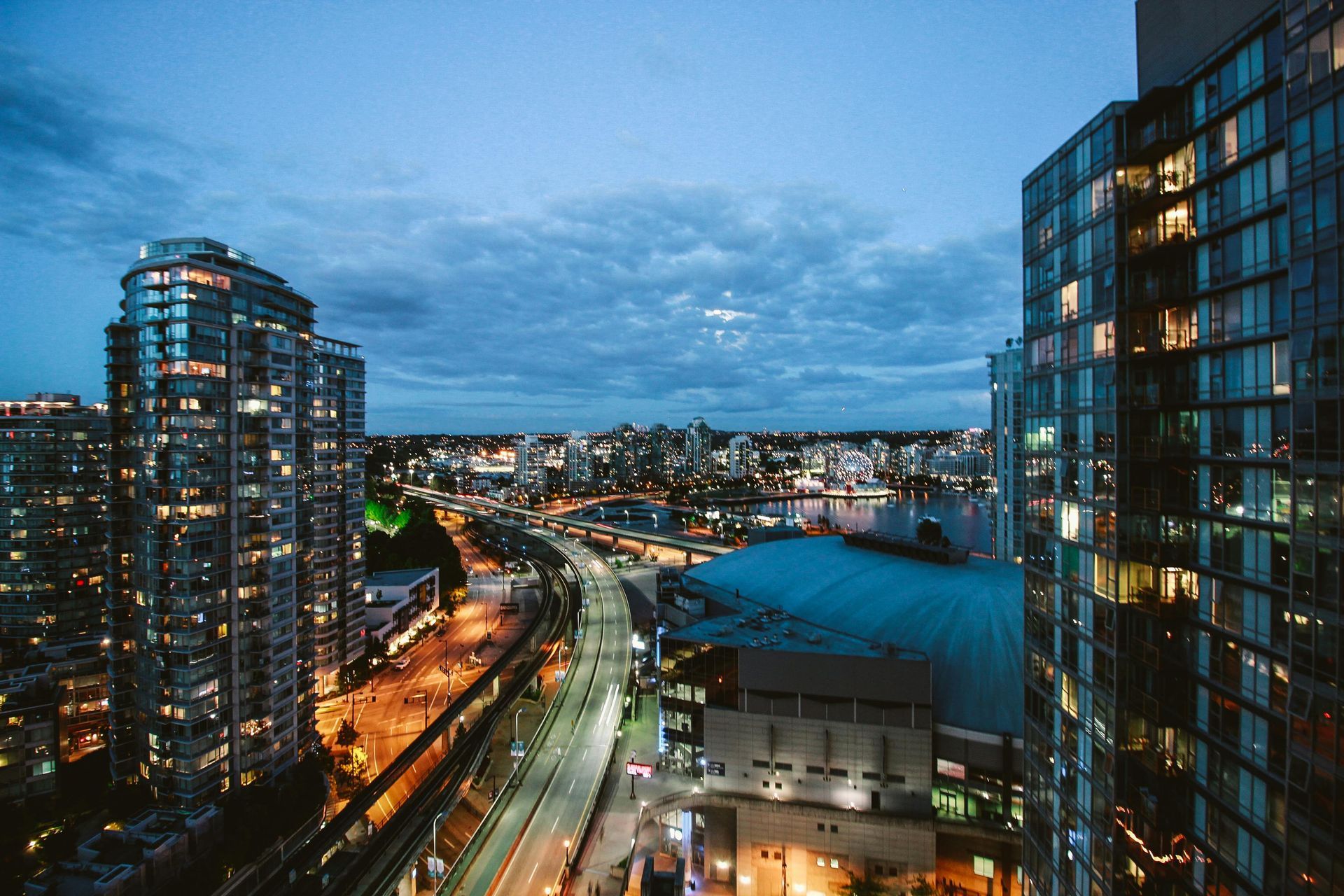 City skyline at dusk with lit buildings, highway, and water under a cloudy blue sky.