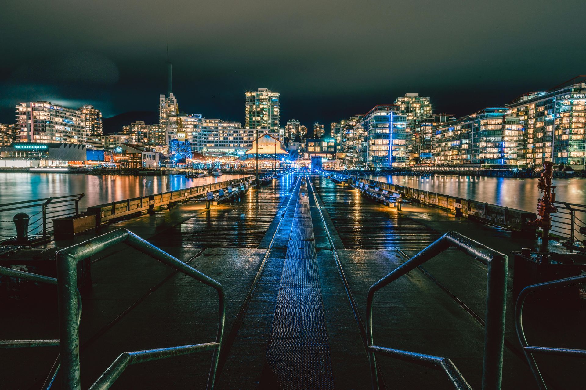 Nighttime view of city lights reflecting on water from a wooden pier, with illuminated buildings in background.