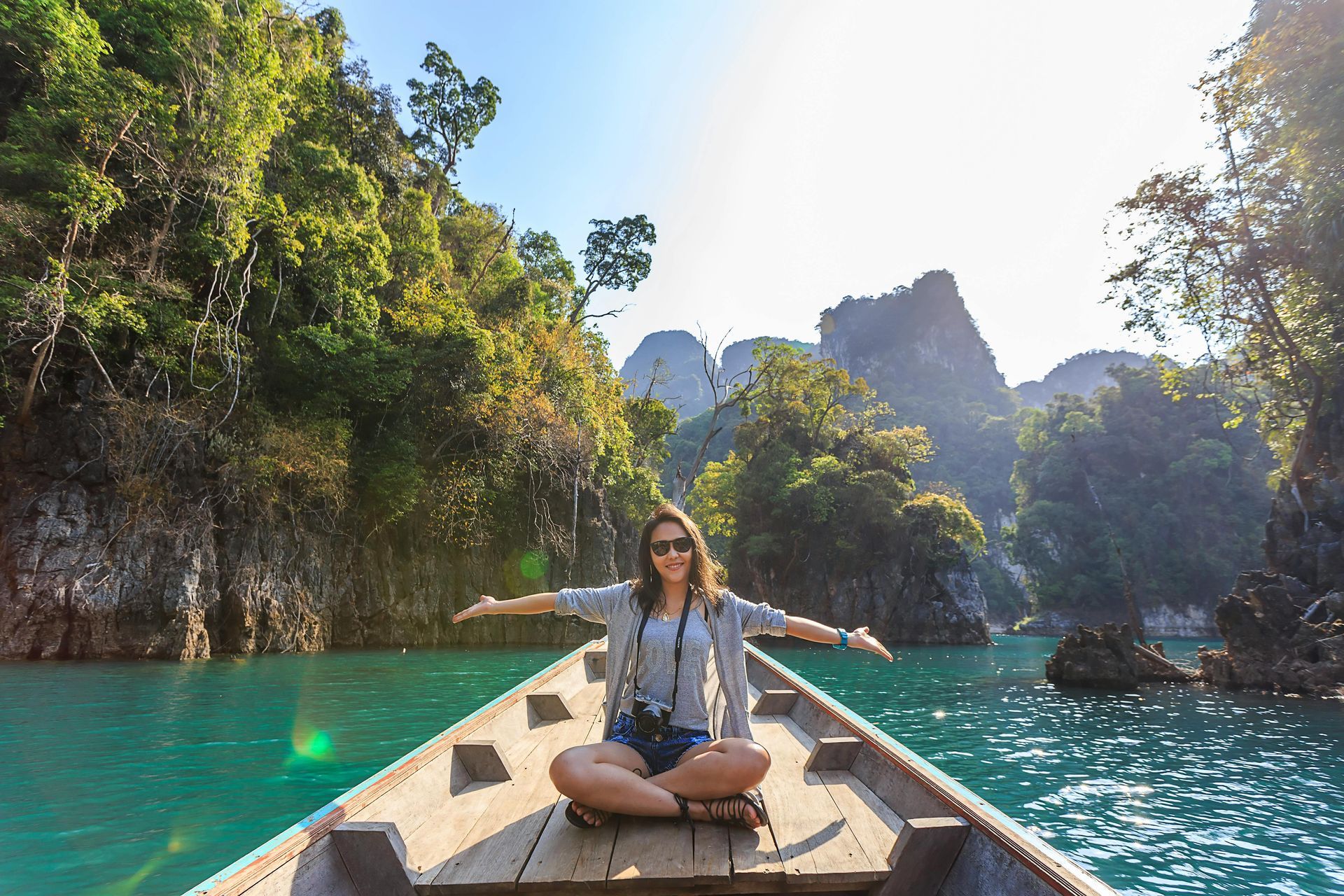 A woman sitting on a boat with arms spread wide as the boat navigates a lagoon in Thailand with greenery on sides of cliffs as it navigates