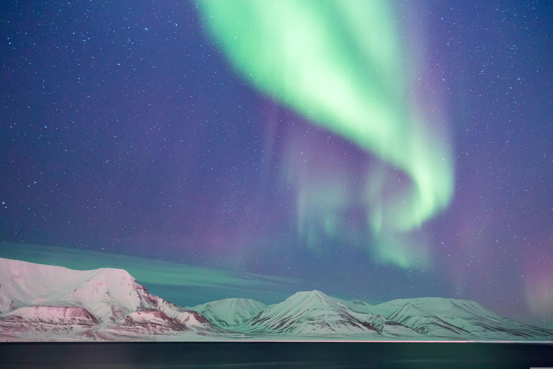 The Aurora Borealis overtop snow covered mountains viewed from the water.
