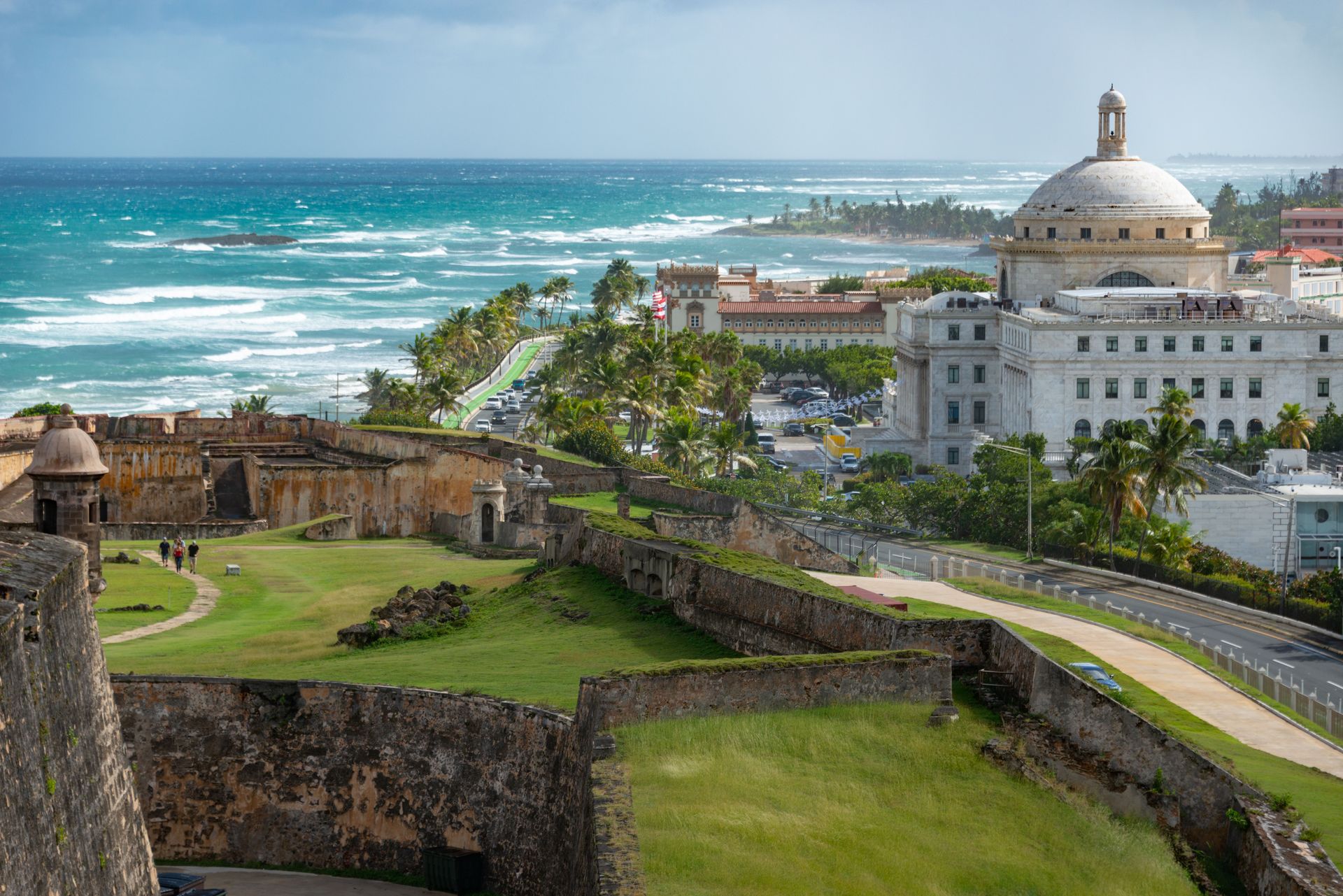 Coastal fortress overlooking turquoise ocean, grassy walls, and a white domed building under blue sky