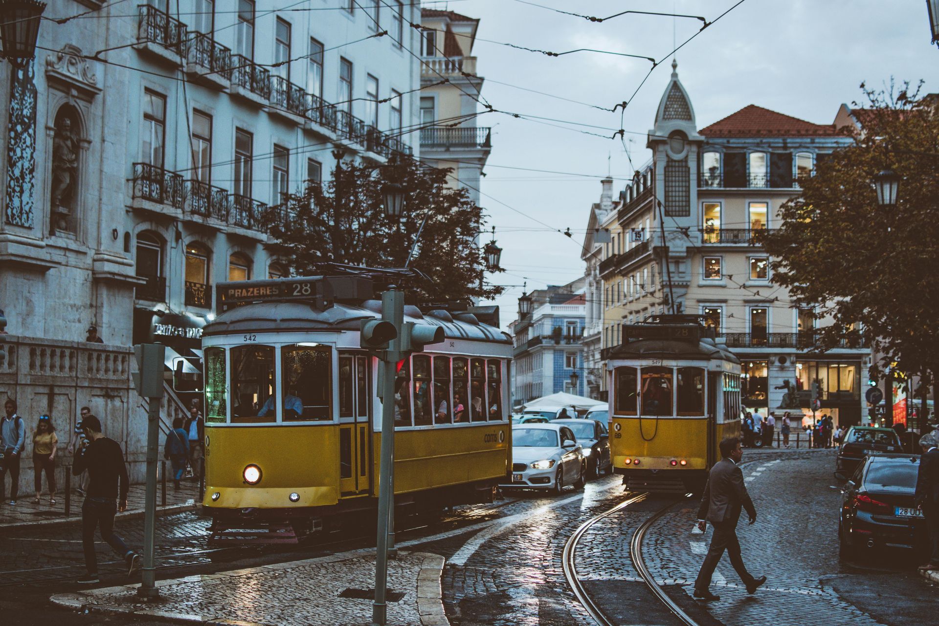 Yellow trams travel on a cobblestone street in Lisbon, Portugal. People walk nearby, buildings in the background.