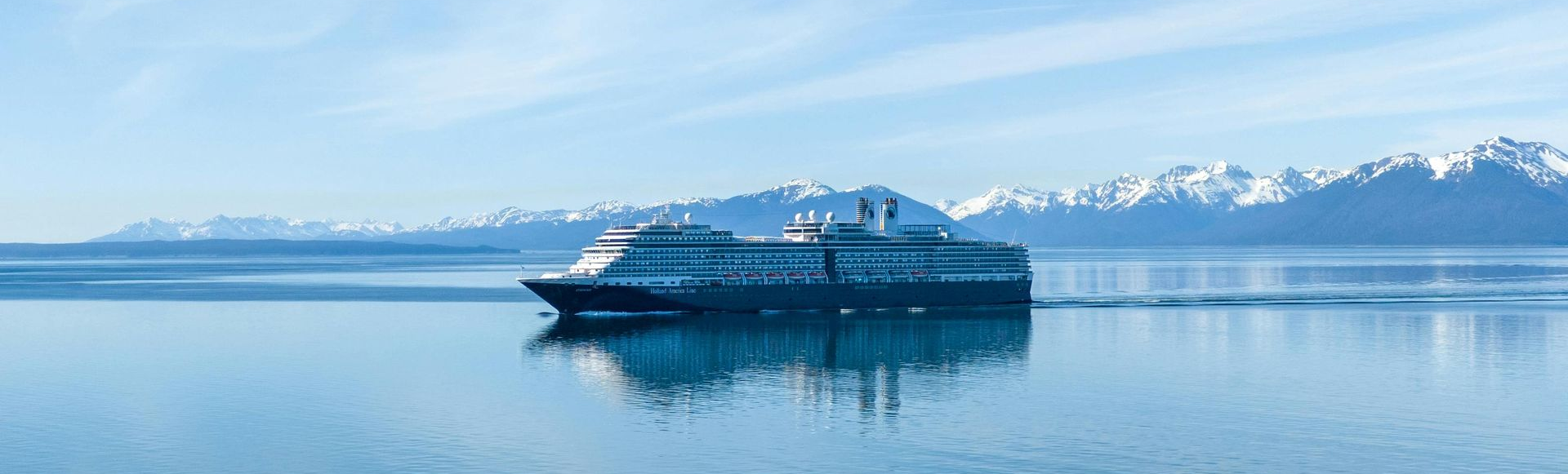 A cruise ship sails on calm water, with snow-covered mountains in the background under a blue sky.