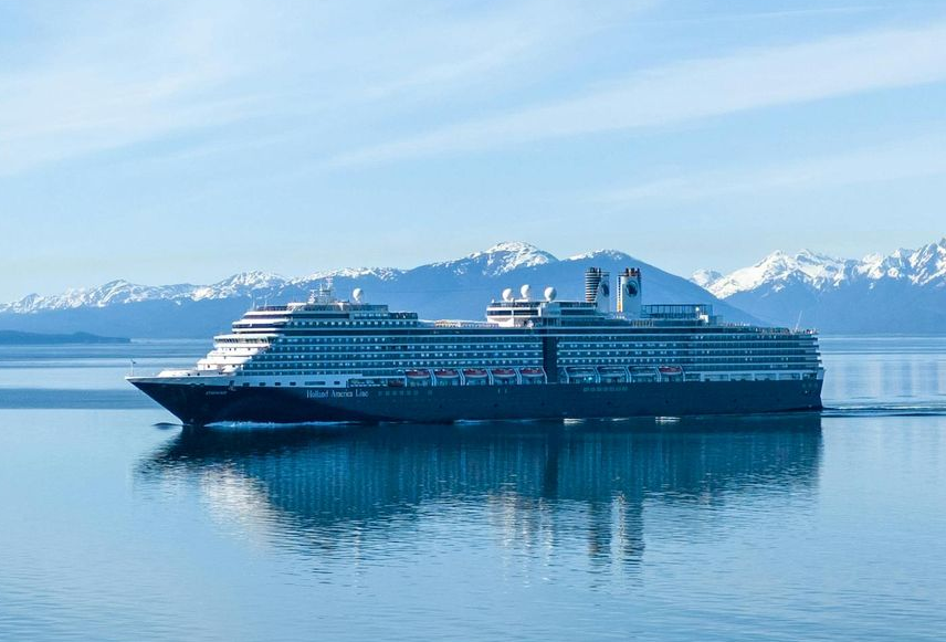 A Holland America Line cruise ship sails through the icy waters of Alaska, surrounded by snow-capped mountains and dramatic coastal cliffs under a clear blue sky.