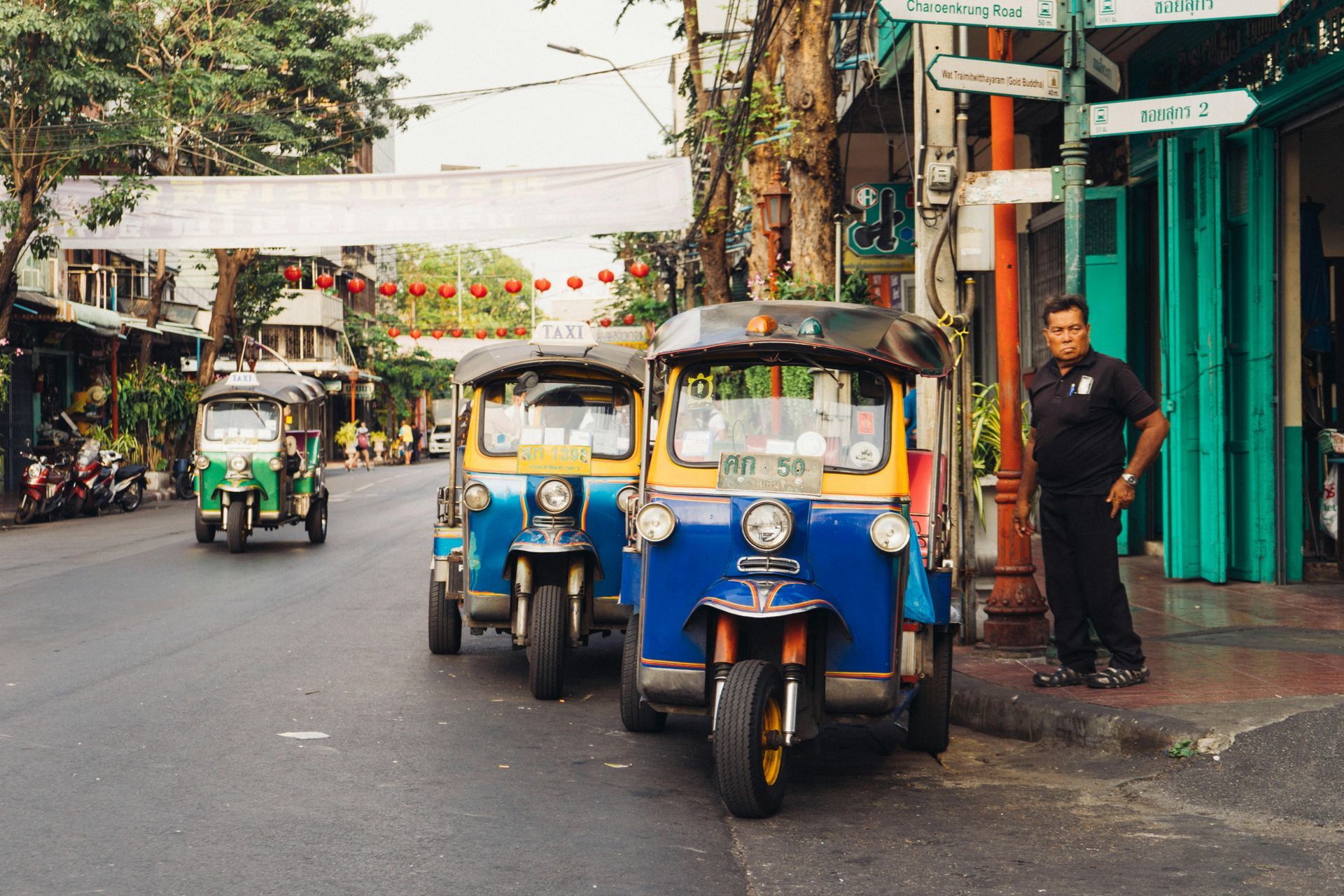 Tuk-tuks lined up on a street in Thailand. A man stands nearby.