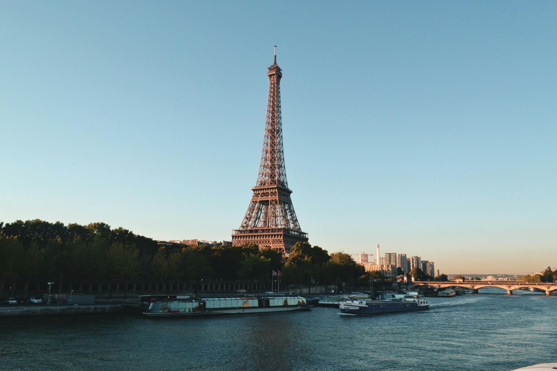 Eiffel Tower in Paris, France, over the Seine River. Blue sky, boats, and lush trees.