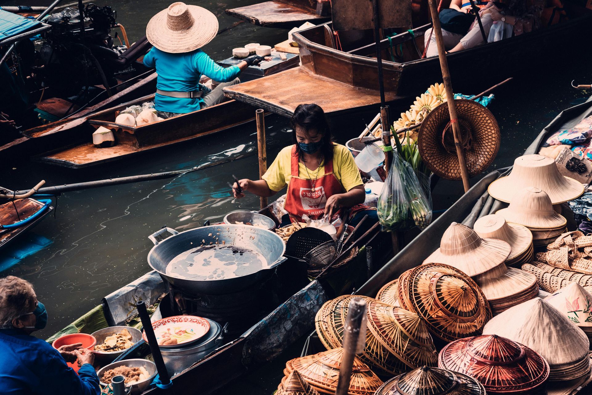 Floating market in Thailand: Vendors cooking food on boats, selling wares; people in hats.