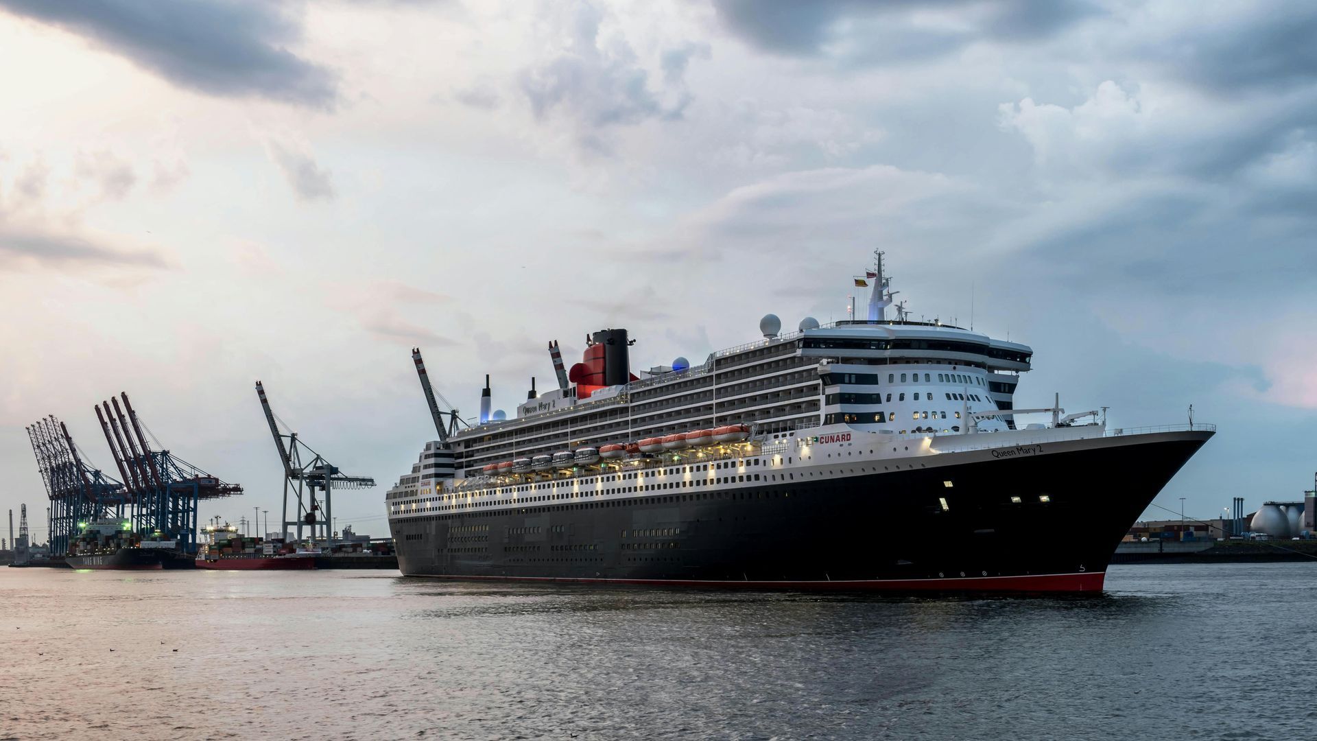 Large cruise ship in a harbor with cranes, black hull, red trim, and white superstructure under a cloudy sky.