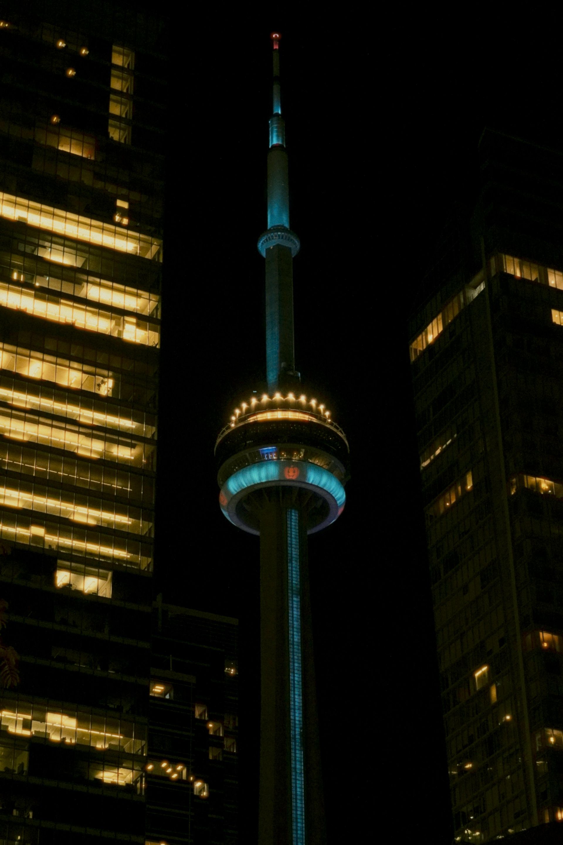 CN Tower illuminated at night, flanked by skyscrapers. Blue, white, and gold lights.