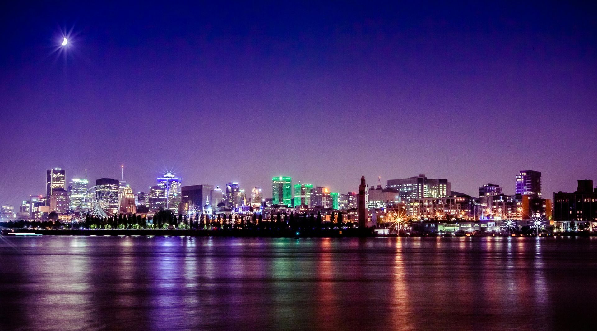 Nighttime cityscape with illuminated buildings reflected in calm water under a dark purple sky.