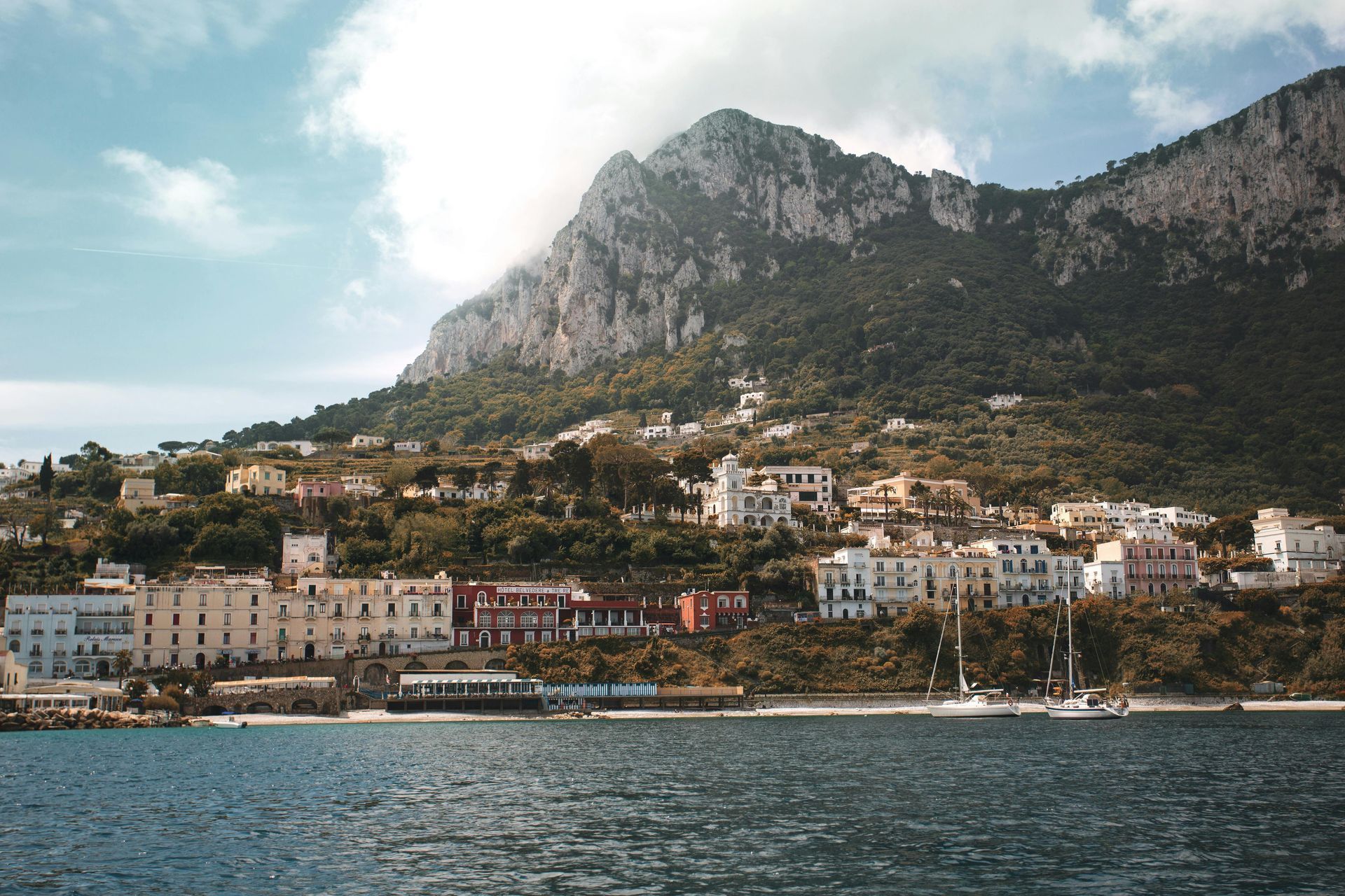 Coastal town nestled at the foot of a mountain. Buildings line the shore; boats are in the water.